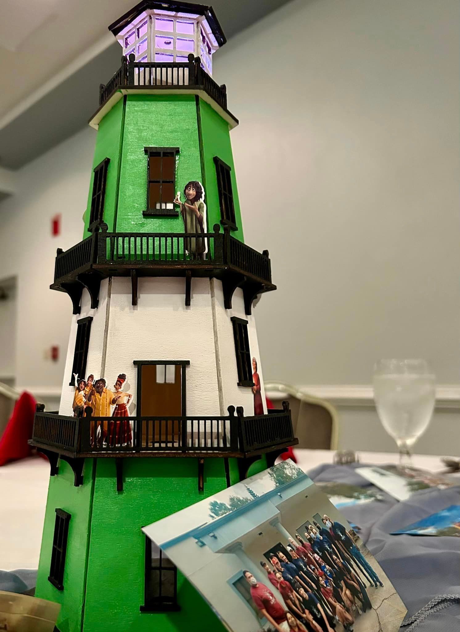 Green and white toy lighthouse with figures on balconies, a lit top, and photos on a table.
