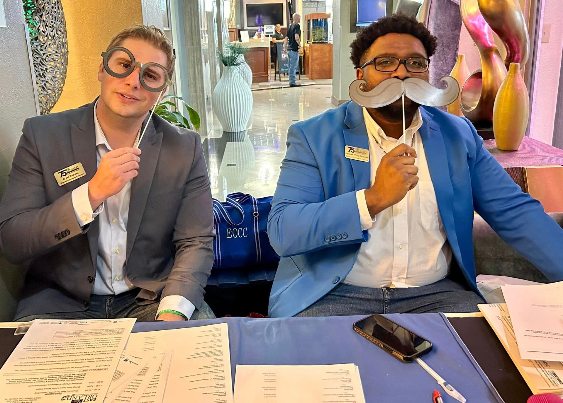 Two people at a desk wearing novelty glasses and a mustache. They are smiling.