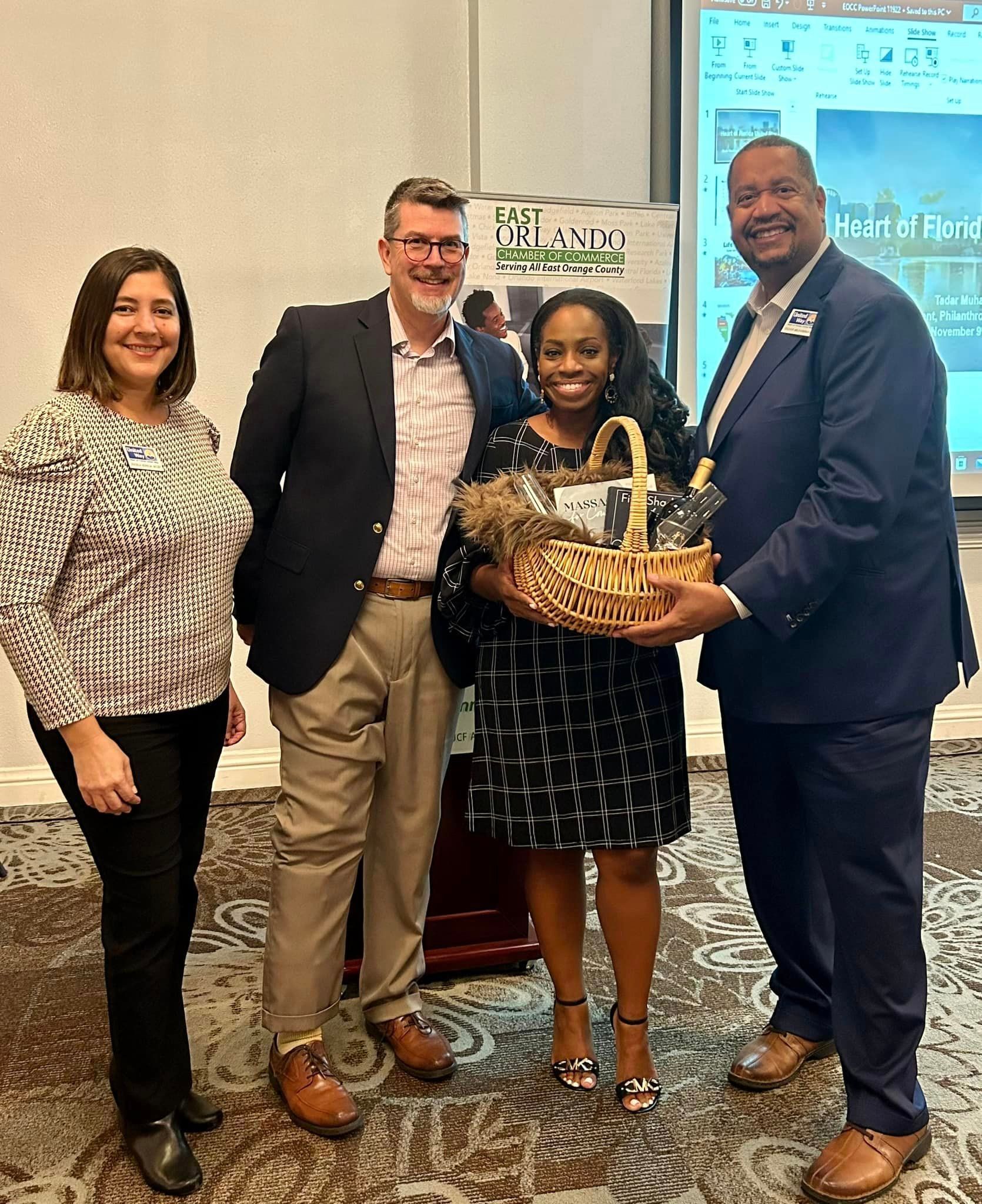 Four people stand with a gift basket; two in suits, one in patterned dress.