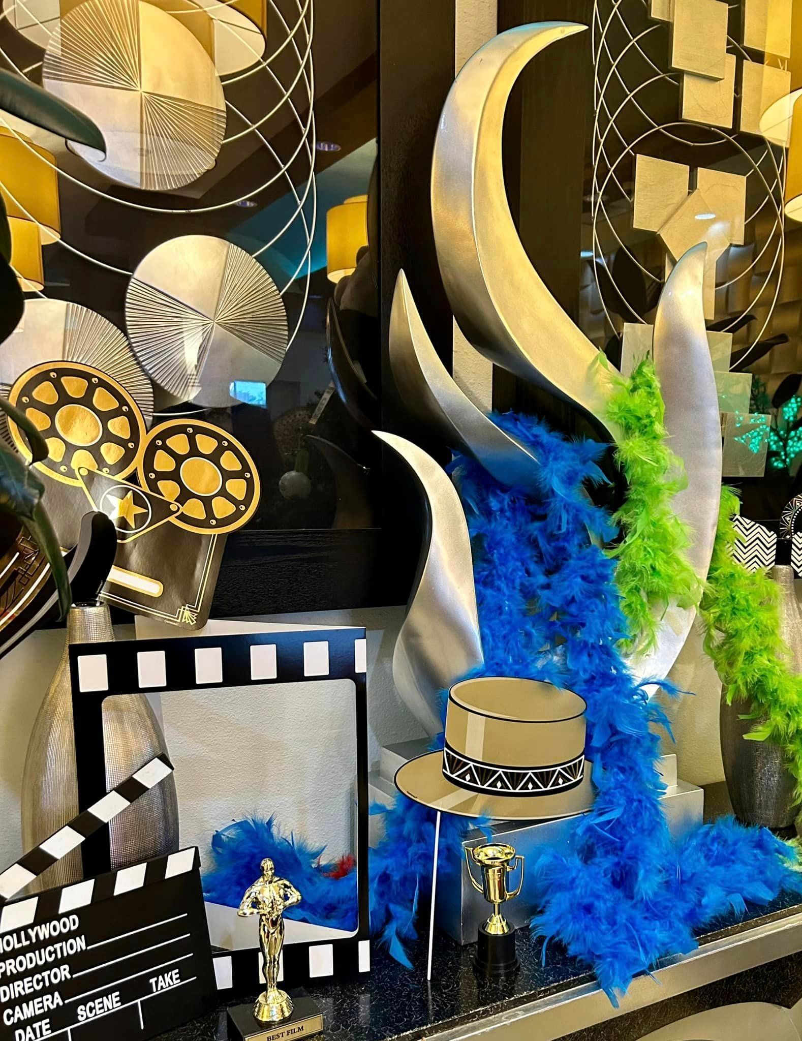 Hollywood-themed display with silver sculptures, a clapperboard, blue boa, and Oscar statues.