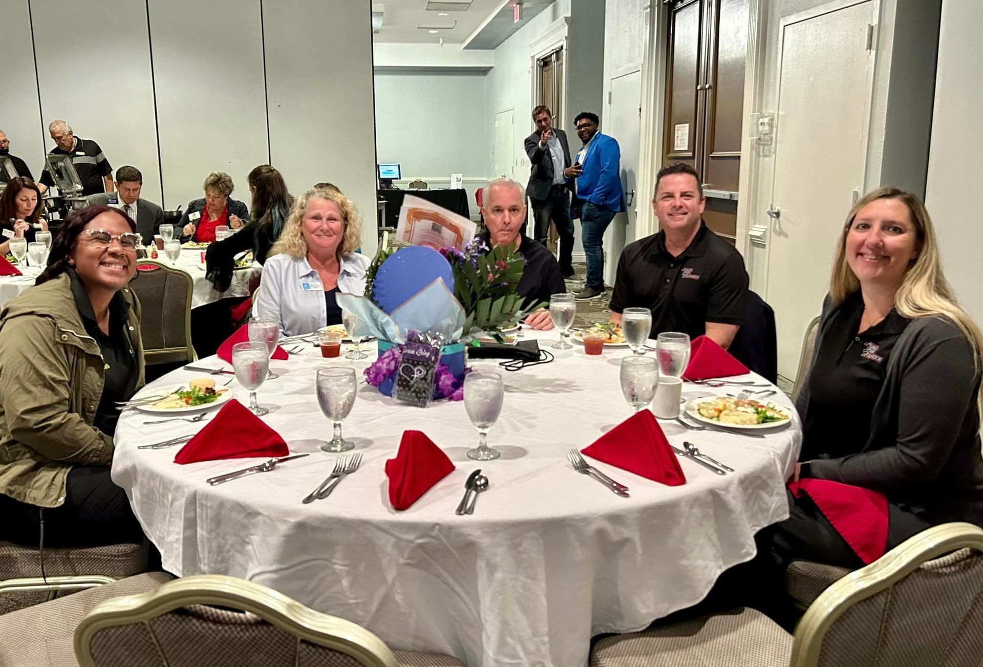 People seated around a table at an event. A floral centerpiece with red napkins, and water glasses are on the table.