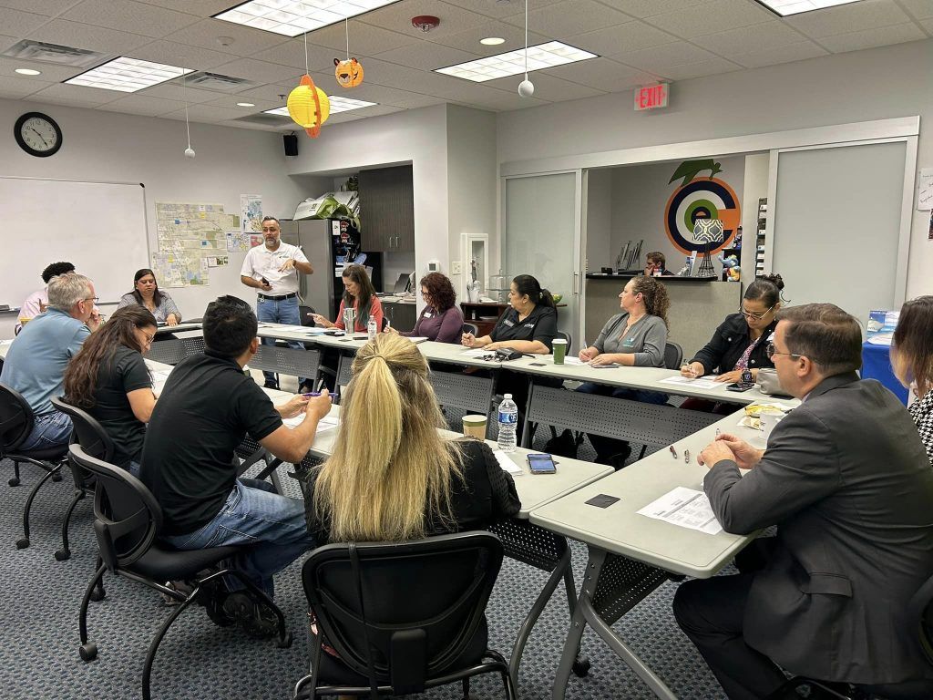 People seated around a table in a meeting, one person standing and speaking, in a brightly lit office.
