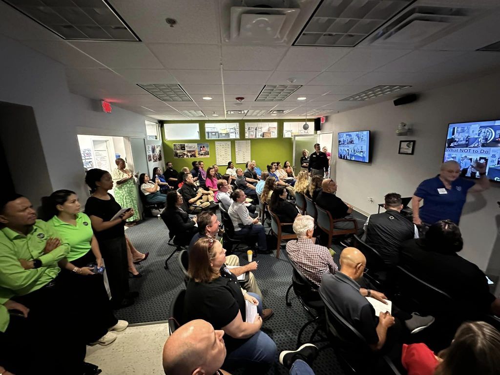 People in a meeting room, listening to a speaker, with screens and a green wall.