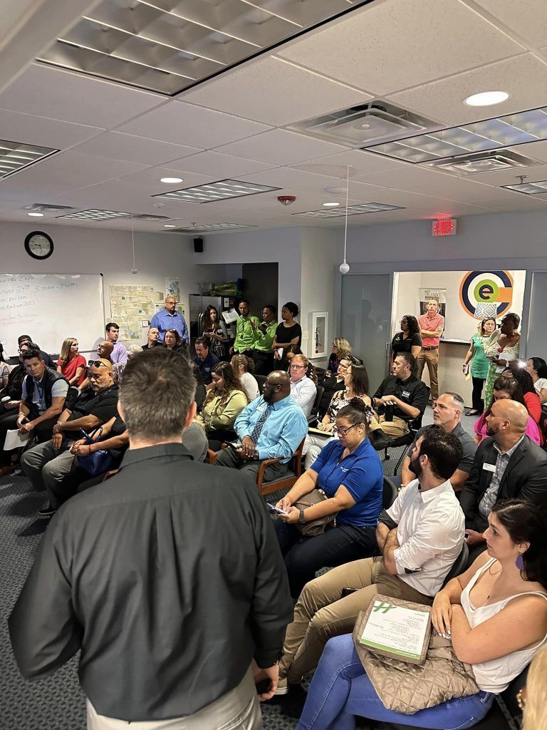 A presentation in a room; a presenter speaks to a seated audience, some looking at a speaker, others away; brightly lit with ceiling panels.