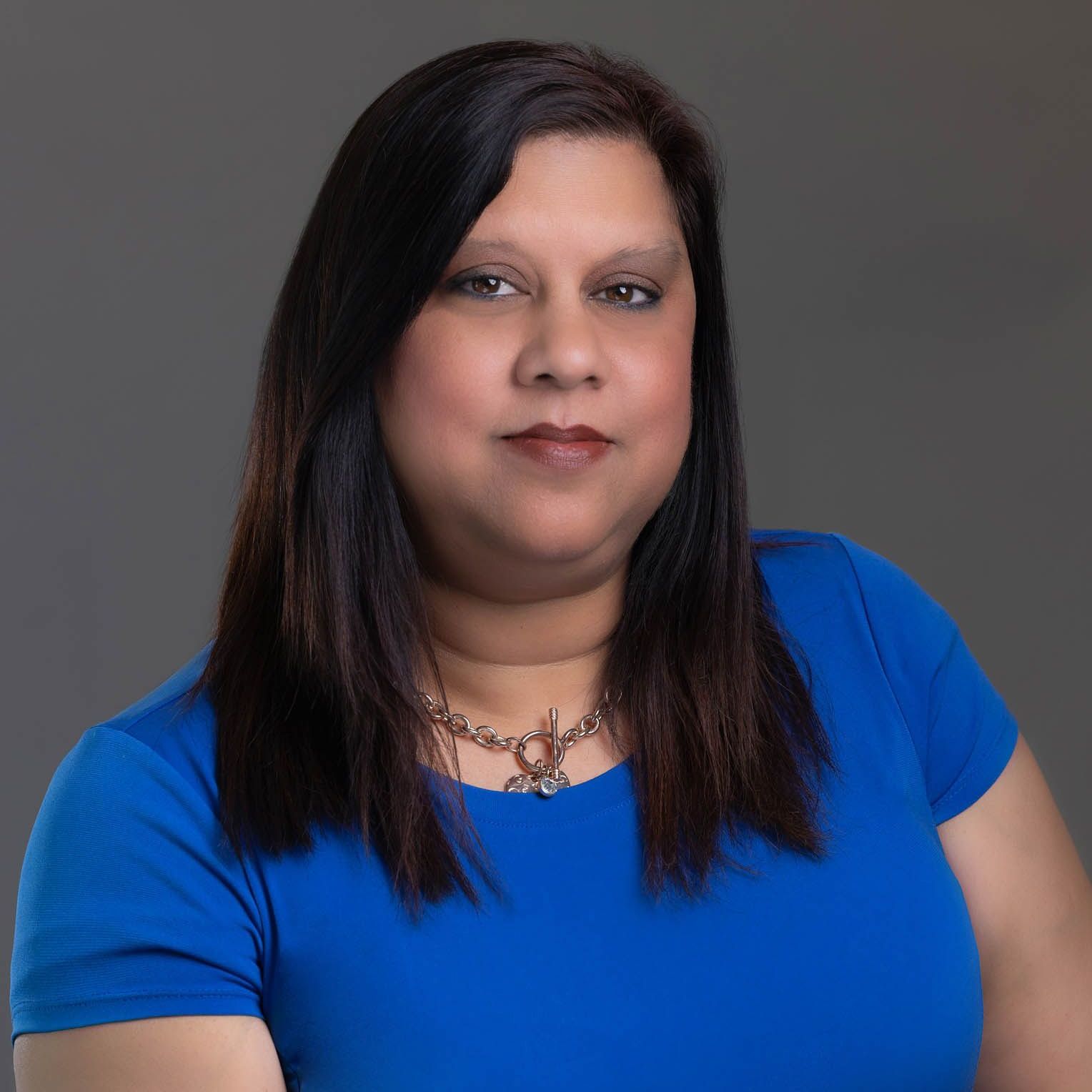 Woman in a blue shirt with dark hair and a necklace, posed against a gray backdrop.