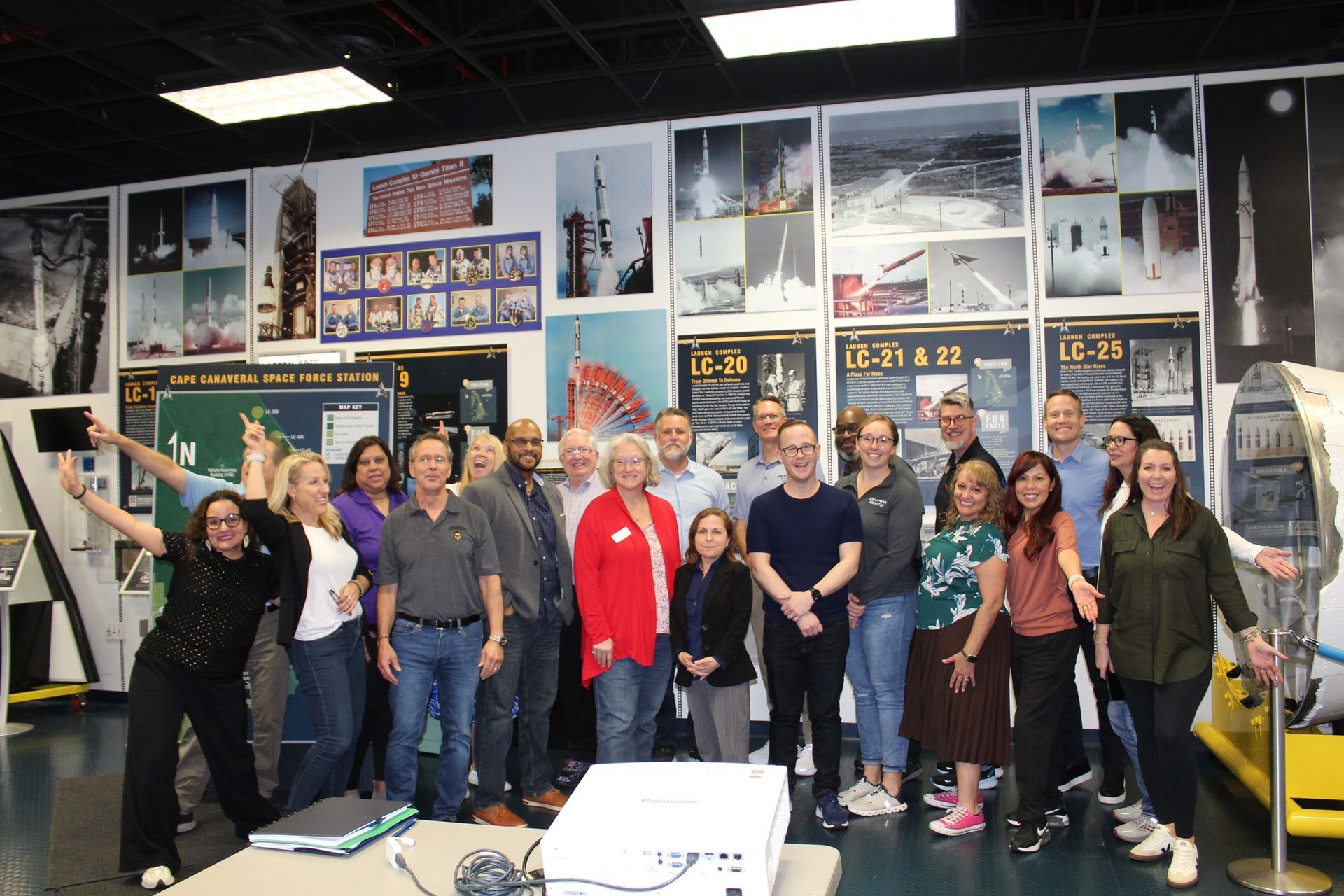 Group of people posing in a room with rocket posters. Some smiling and looking at the camera. A projector is on a table.