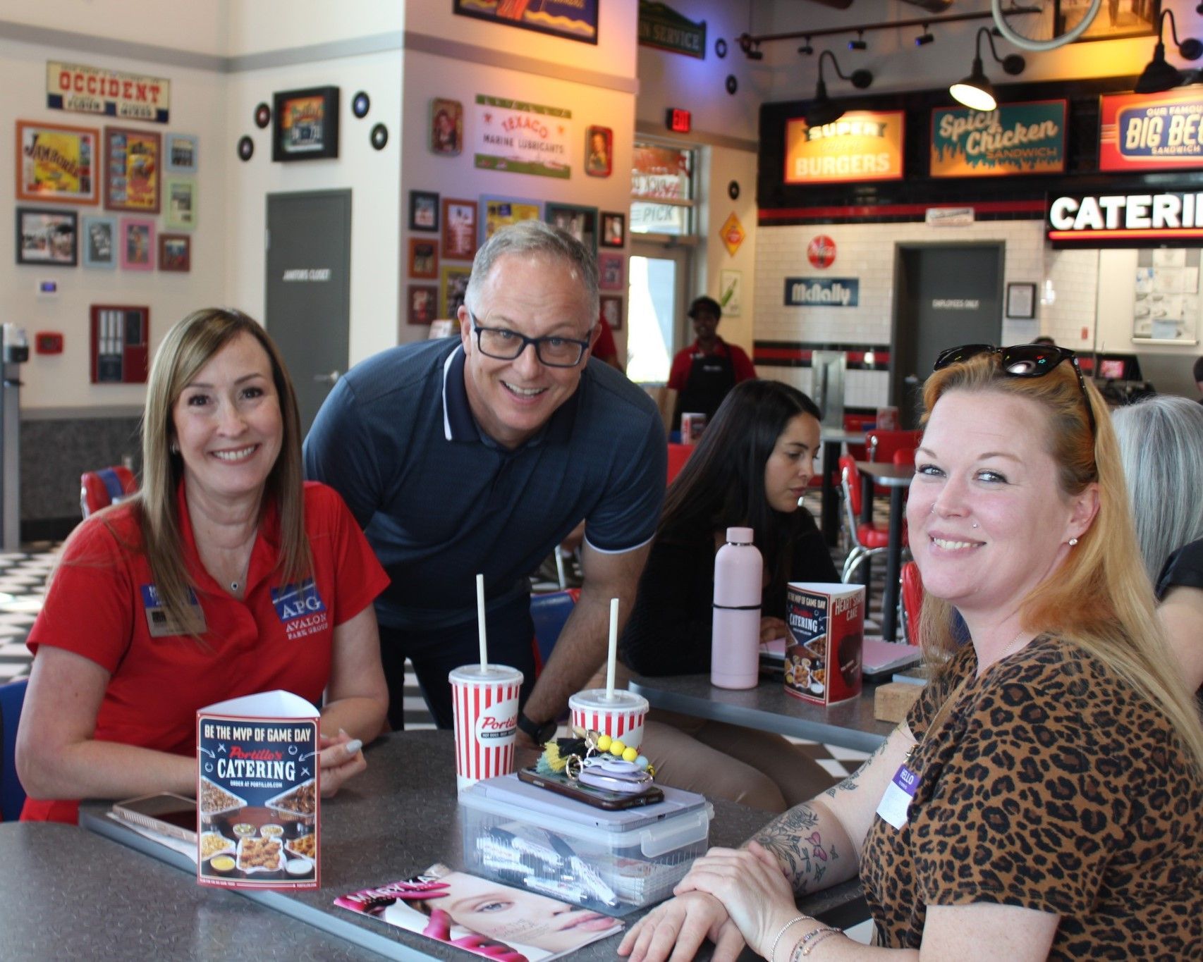 People at a diner table, smiling. One holds a menu, others have drinks. Diner interior with decor.
