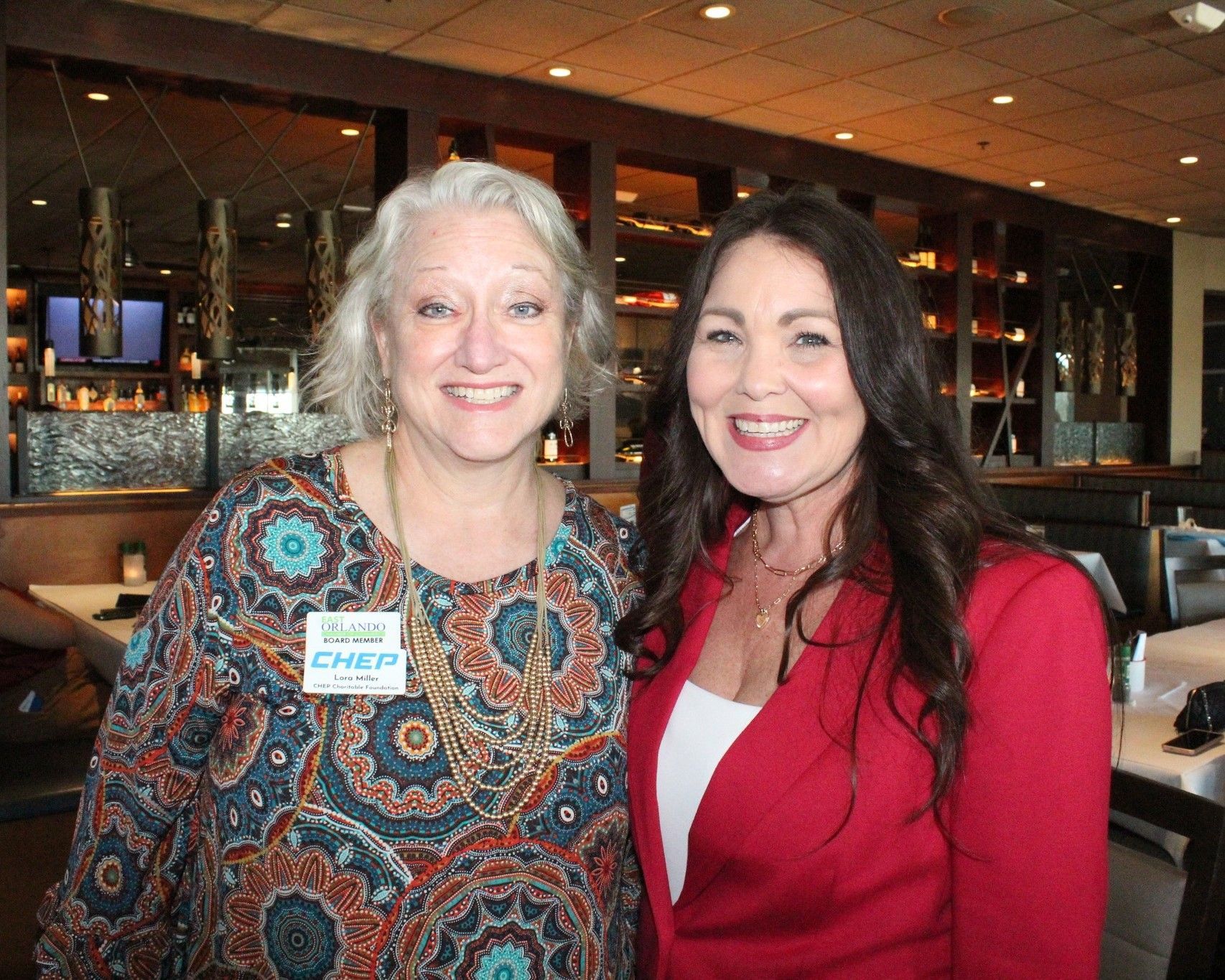 Two women smiling for the camera in a restaurant. One wears a patterned shirt, the other a red blazer.