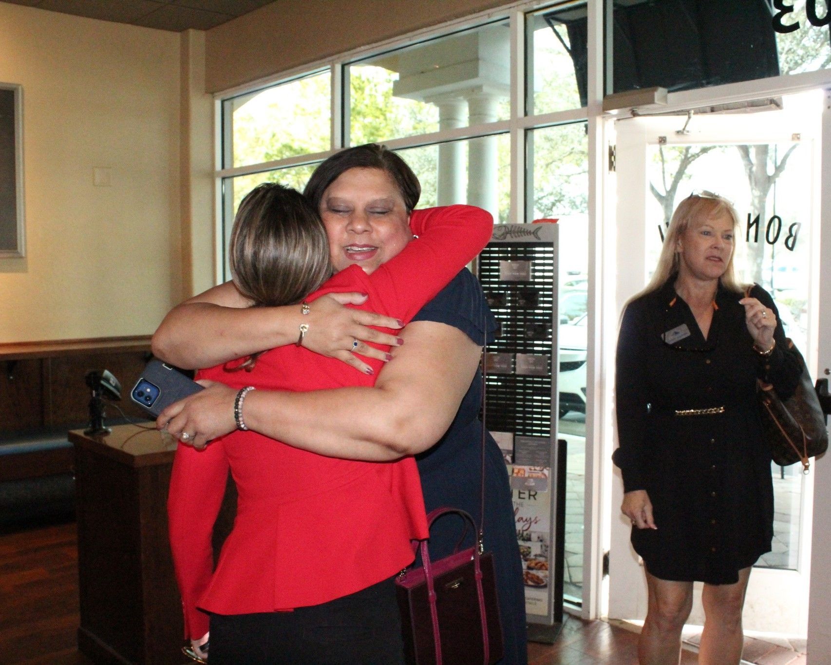 Two women in a warm embrace indoors; one in red jacket, the other in a navy dress. Another woman looks on near an open door.