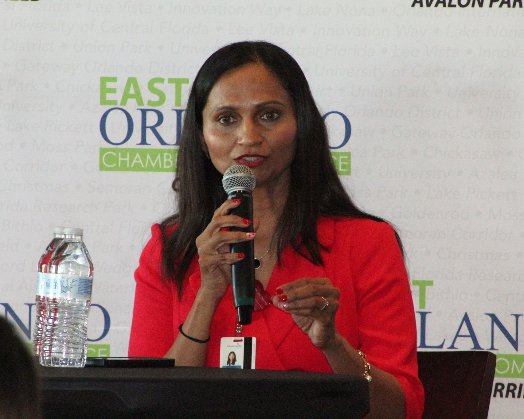 Woman in red blazer speaking into a microphone at an event, East Orlando Chamber logo visible.