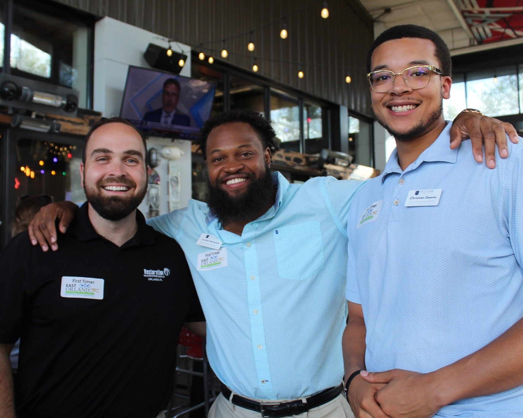 Three men smiling, arm around shoulders. Outdoors. One in blue, one in black, and one in light blue shirt.