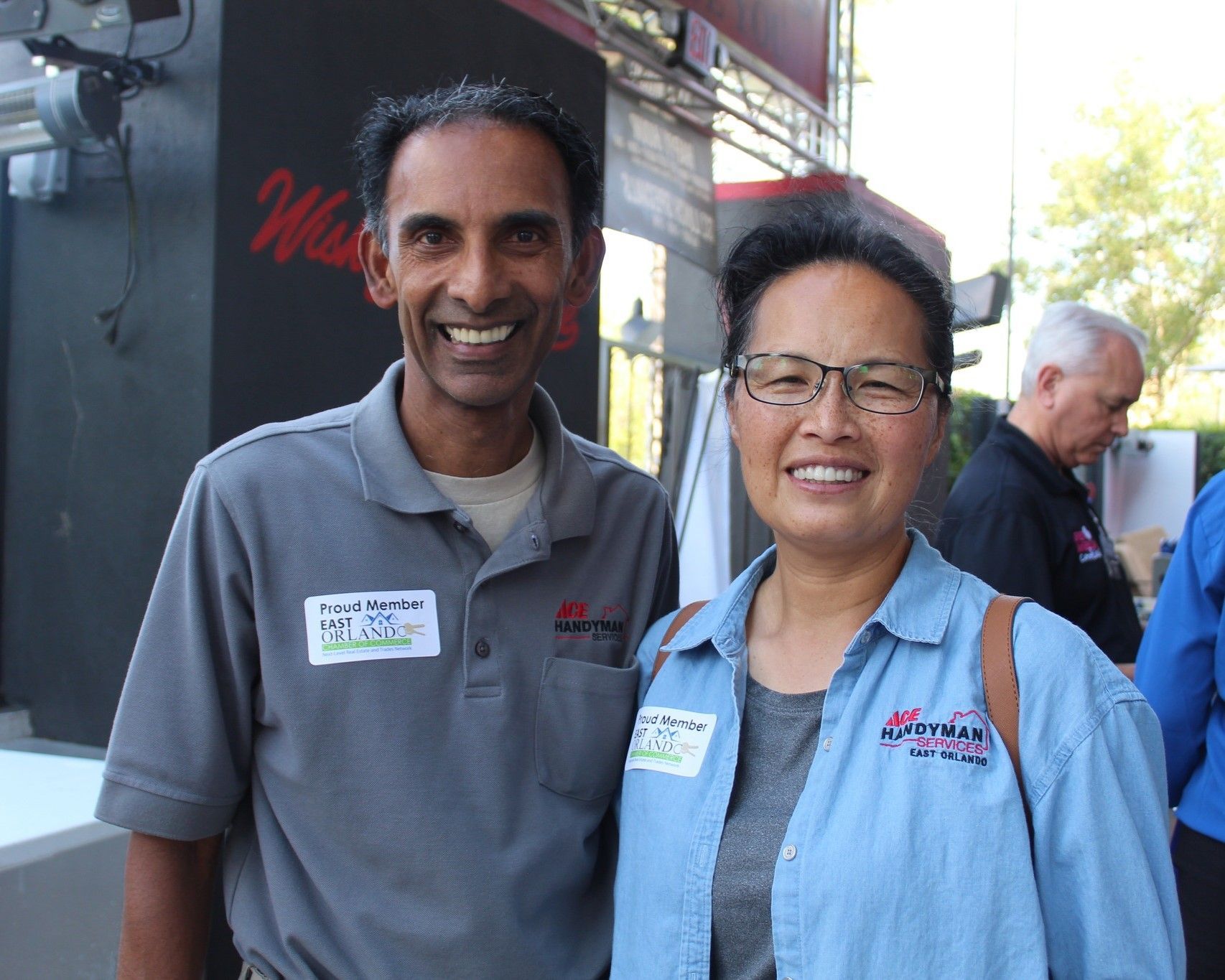 Two people smiling, standing outside a building. Both have name tags and are wearing company shirts.