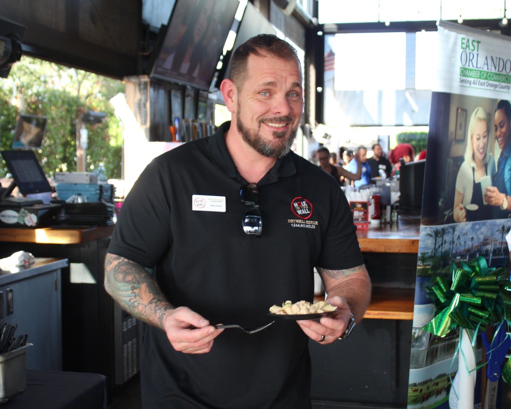 Man holding food at an outdoor event, smiling. Black shirt with logo, tattoo on arm.