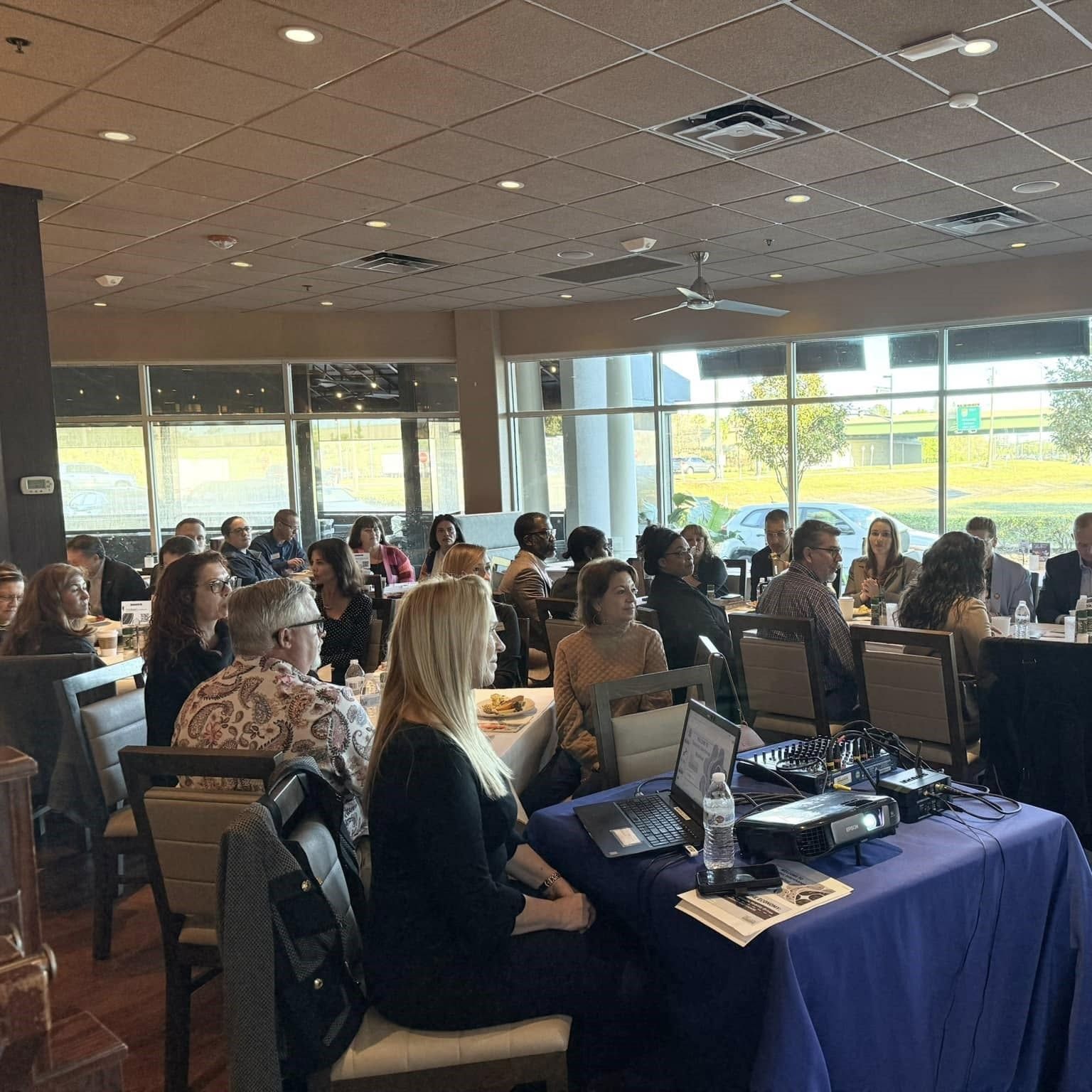 People seated at tables in a room, appearing to be an event or meeting. Sunlight streams in.