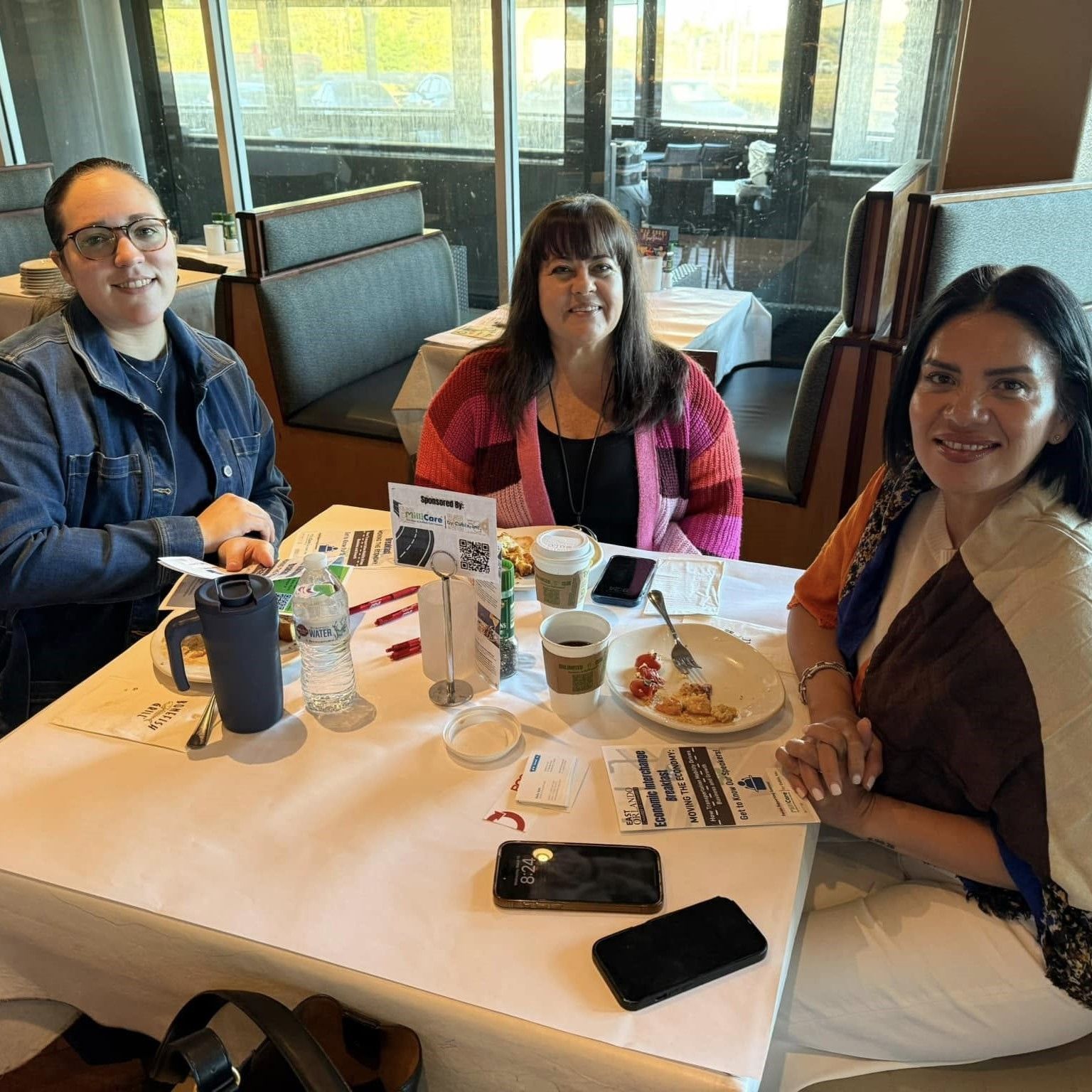 Three people seated at a restaurant table. Two coffee cups, food, and phones visible. Windows and booths in the background.