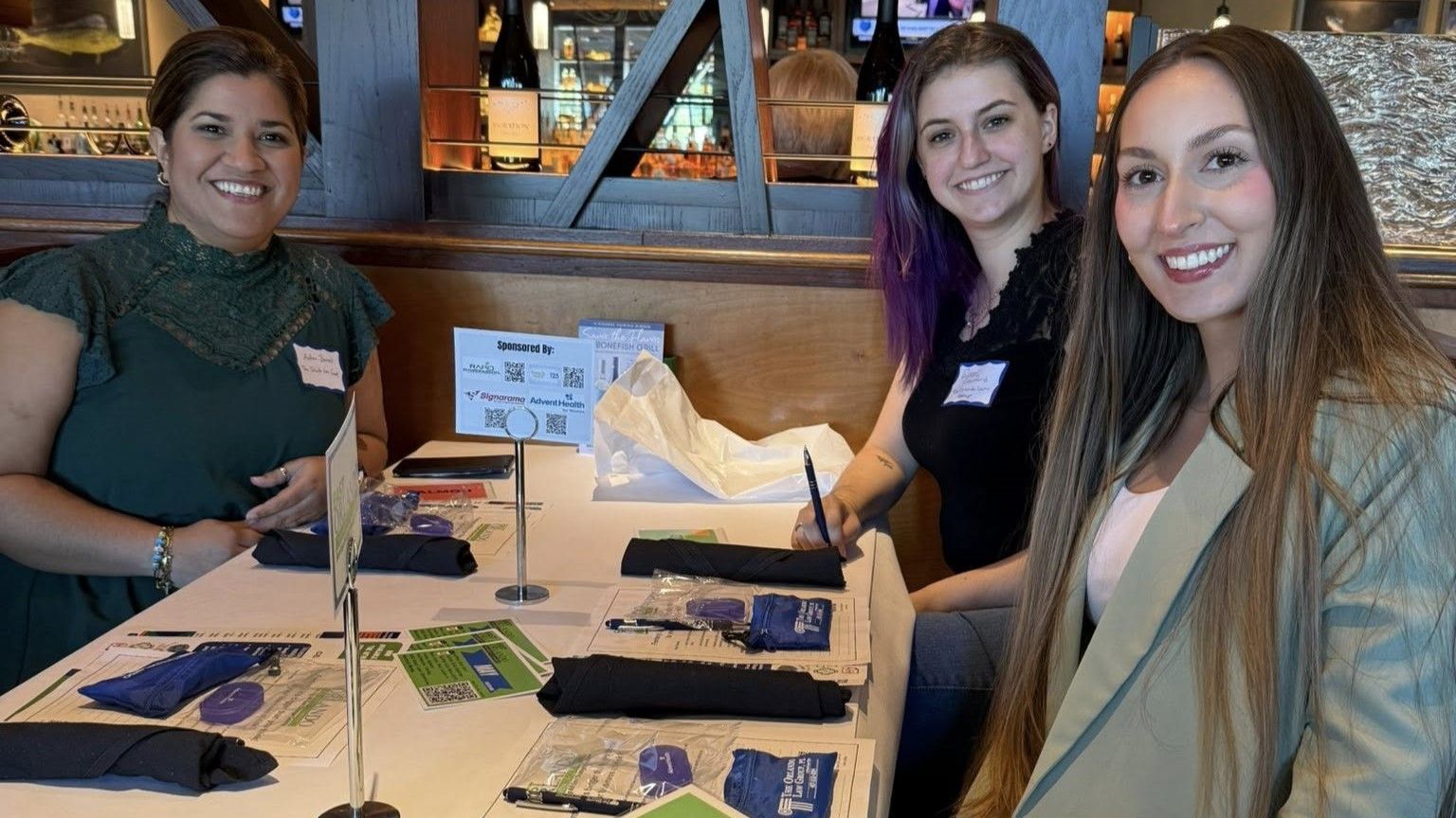Three women smiling at a table, one signing something, inside a restaurant.