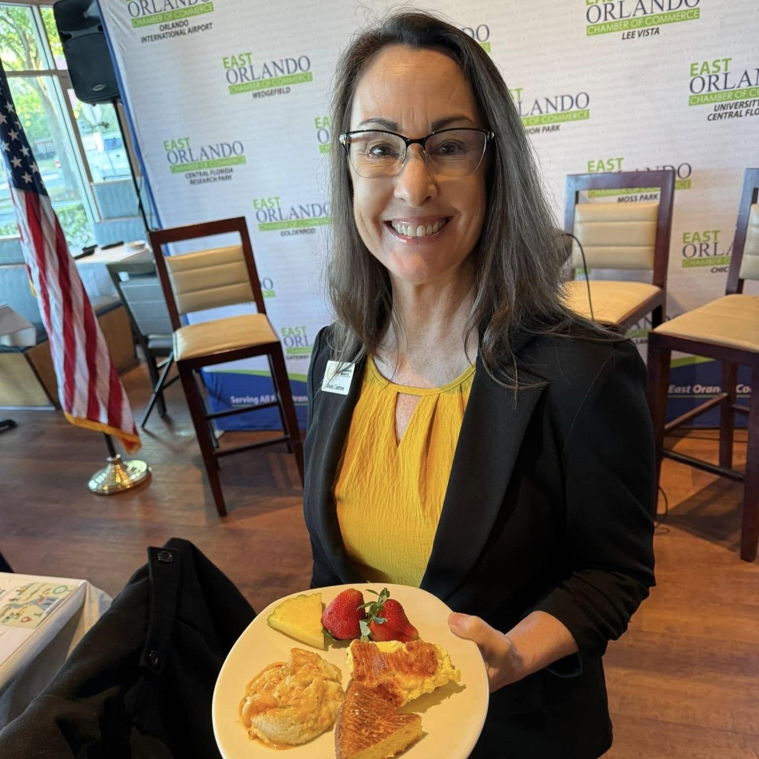 Woman smiles, holding a plate of food. She wears glasses and a black blazer, standing in a conference setting.