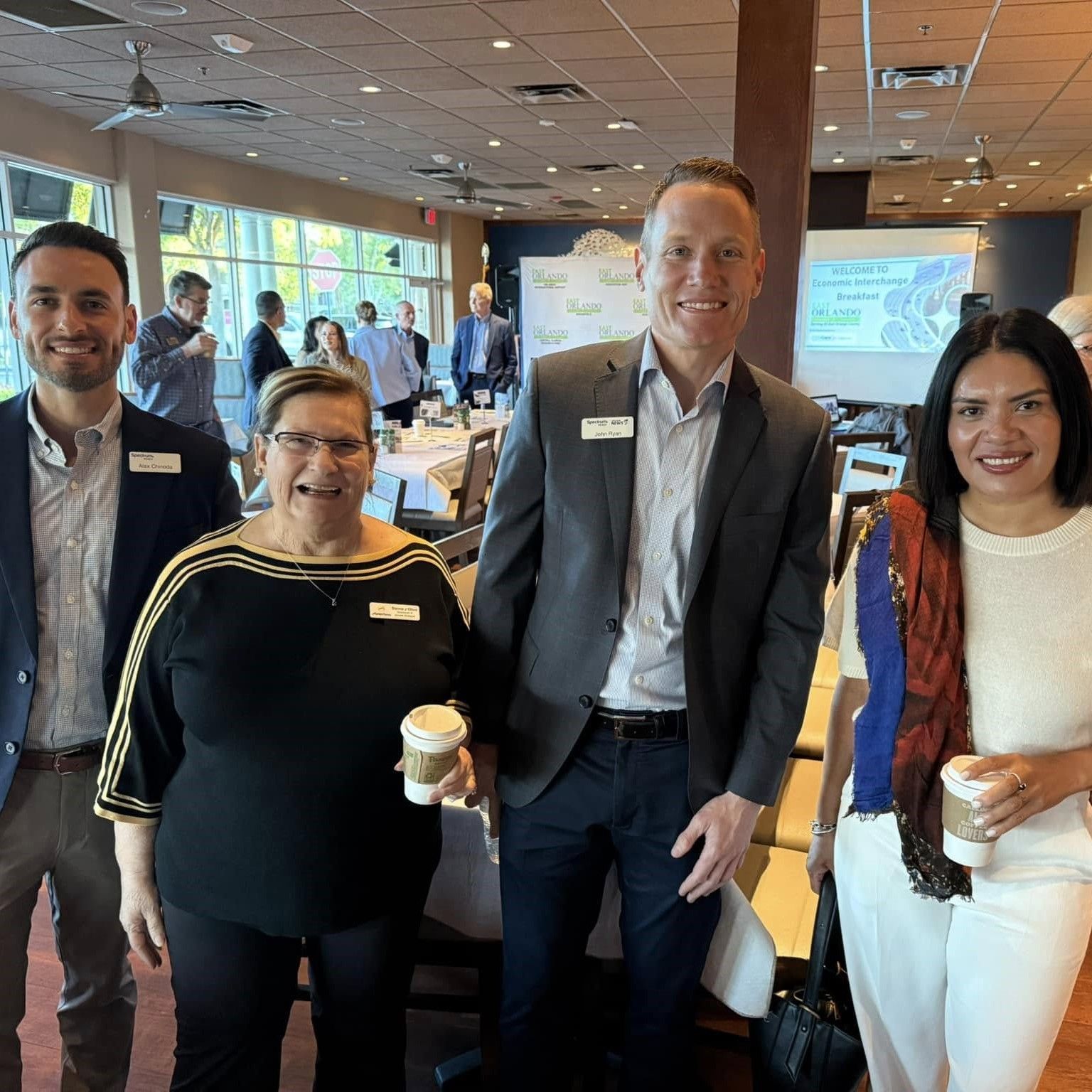 Four people smiling and posing together at an event. A woman holds a coffee cup. Others in the background.