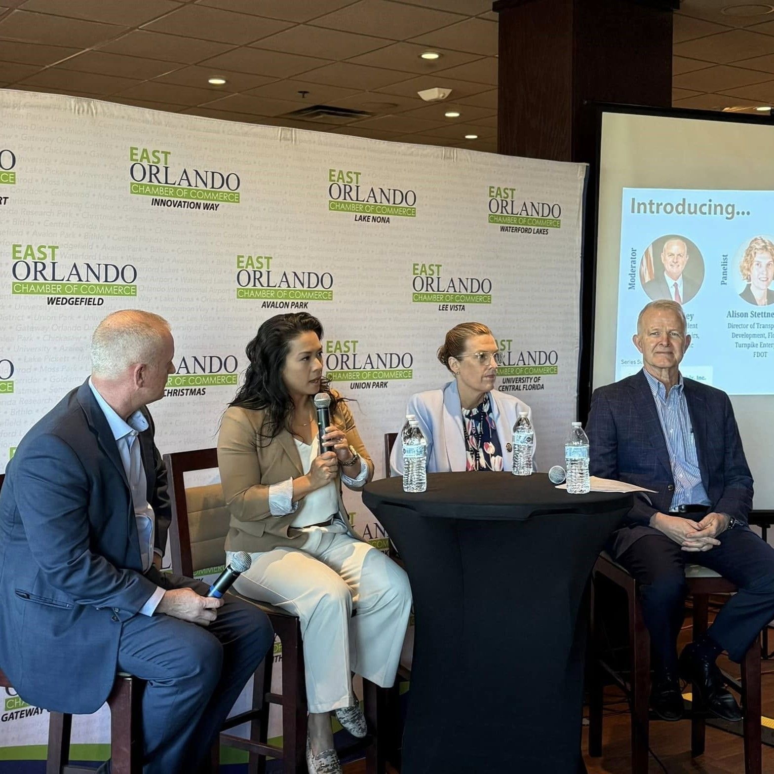 Panel of four people at a table, East Orlando backdrop. Woman speaks into mic; two men, one woman listen.