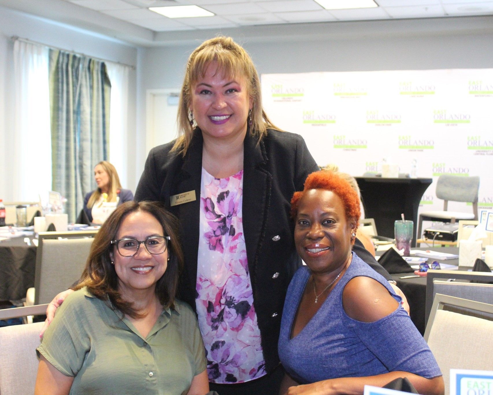 Three women smiling and posing at an event. One in a black blazer, one in a green top, and one in a blue top with bright orange hair.