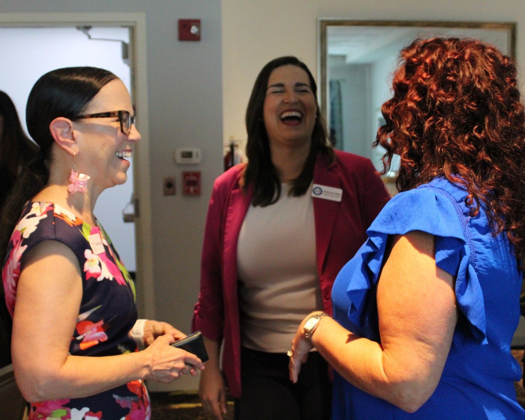 Three women laughing together in a room. One wears a floral dress and glasses, another a pink blazer, and the third a blue top.