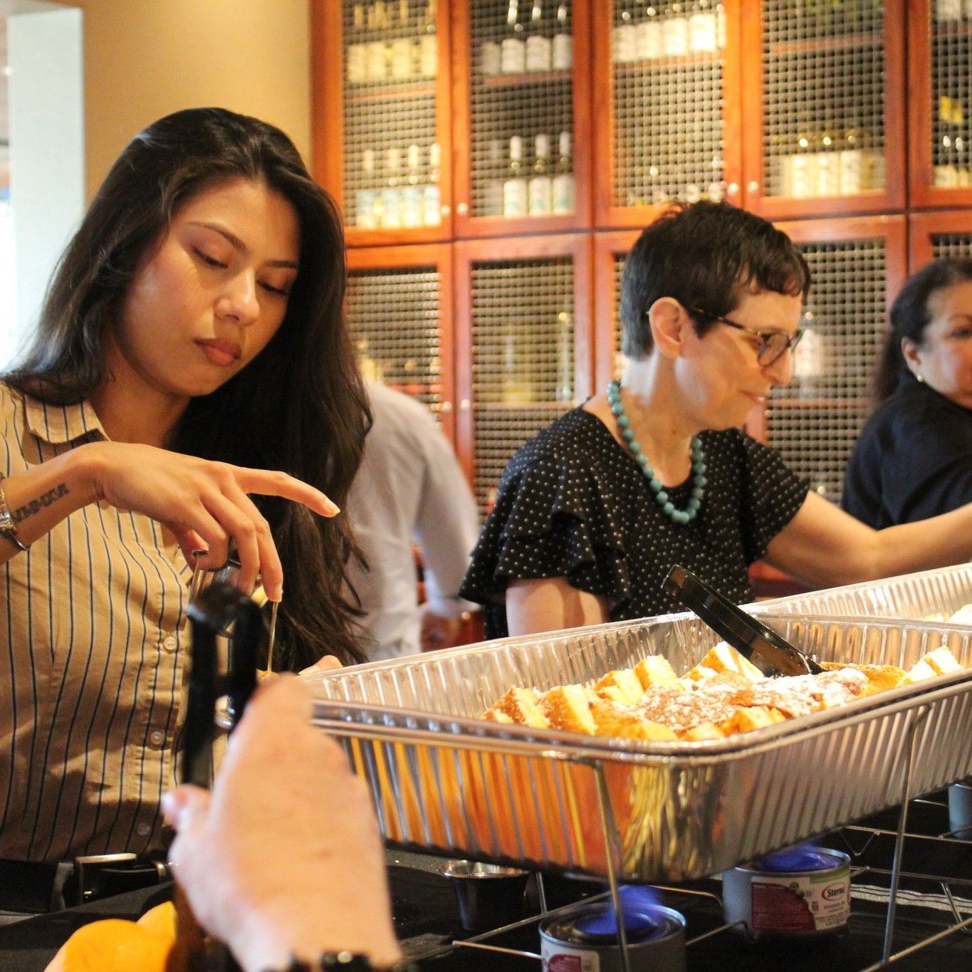 Women serving food from a buffet with a wine rack background. One woman pouring.