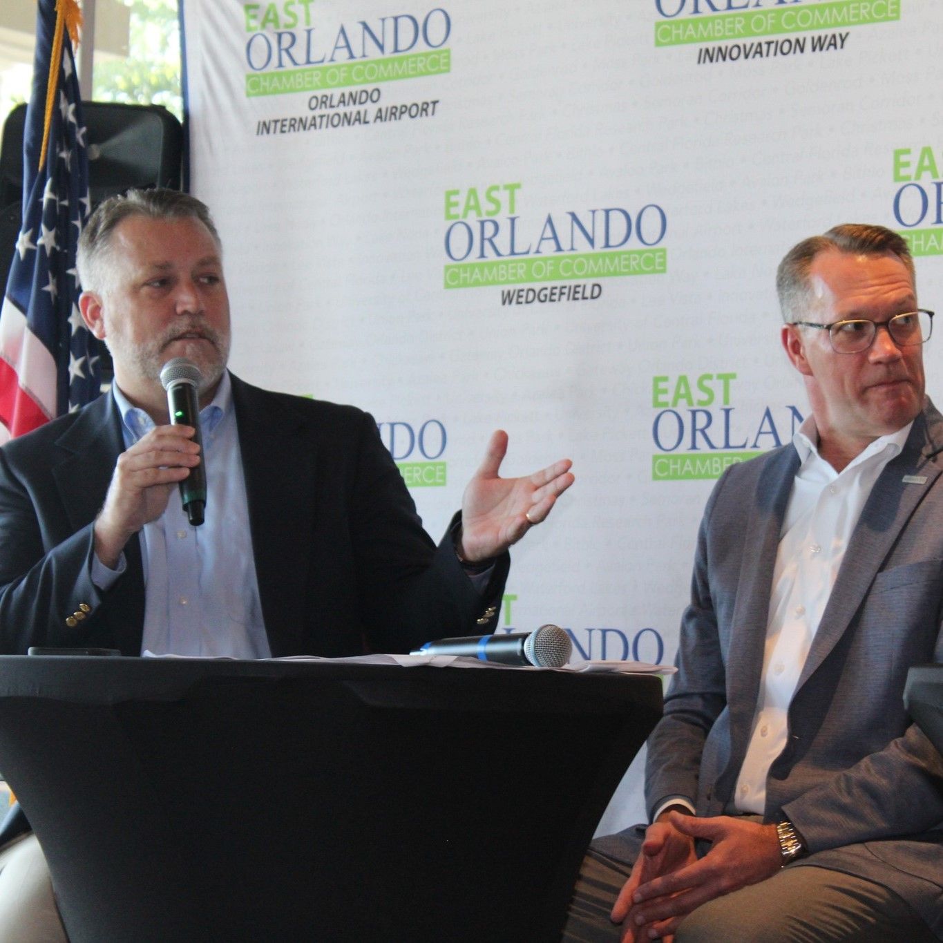 Two men at a panel, one speaking into a microphone. Backdrop has 