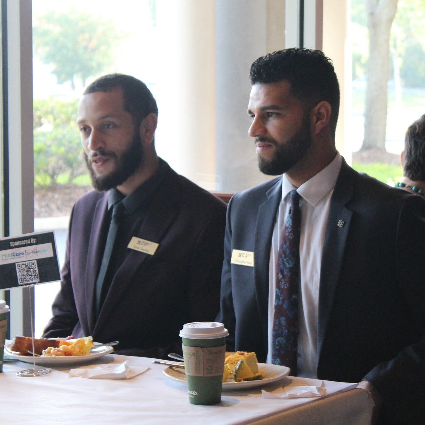 Two men in suits at a table with food, looking toward the right, indoor setting.