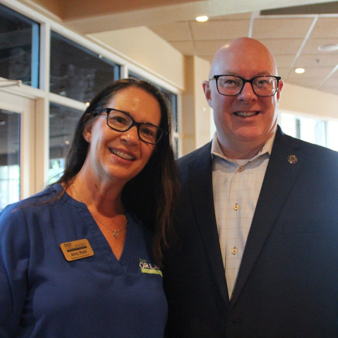 Woman in blue scrubs and man in a suit smiling. Indoors, next to a window.