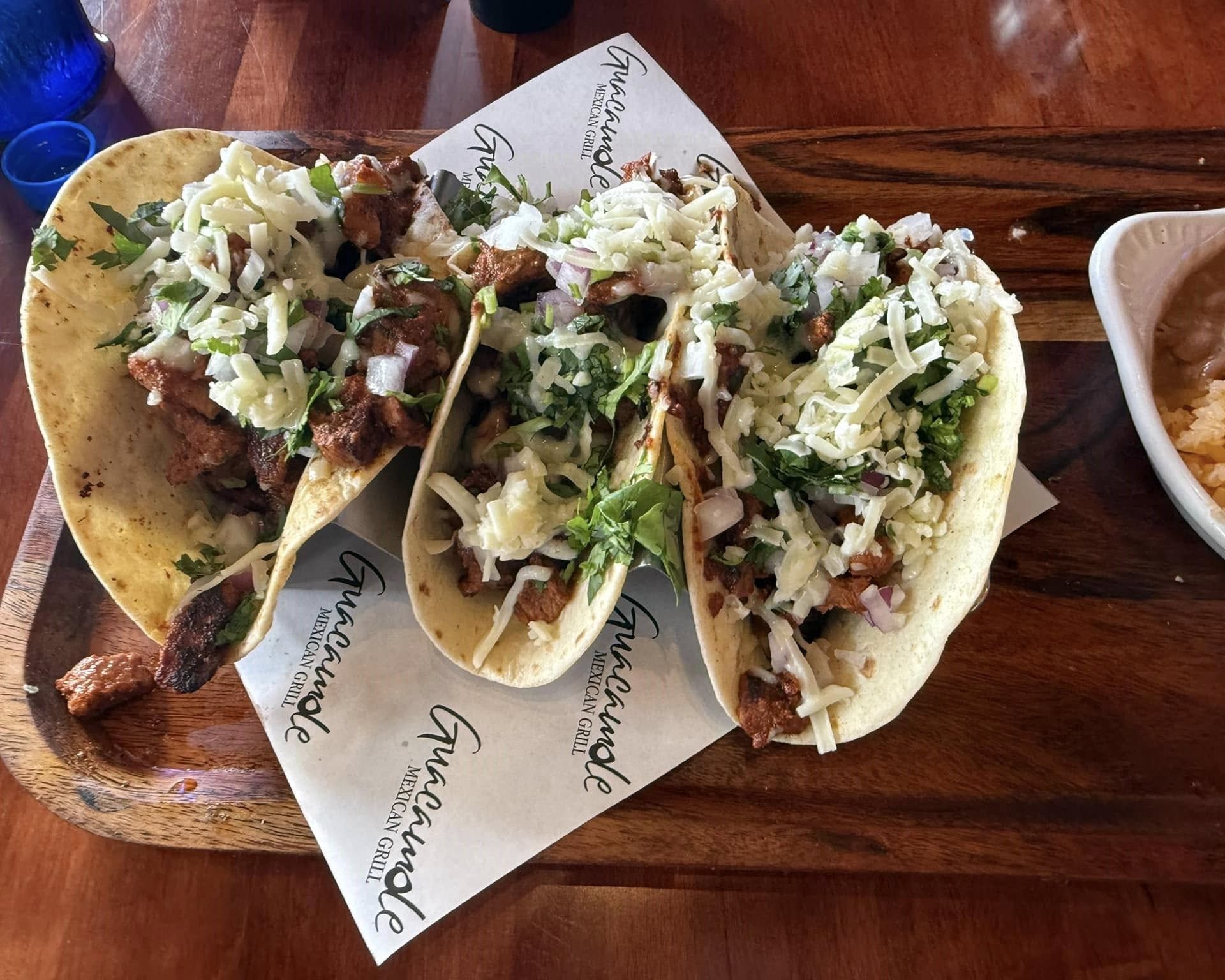 Three tacos with toppings on a wooden tray, next to a small bowl of beans.