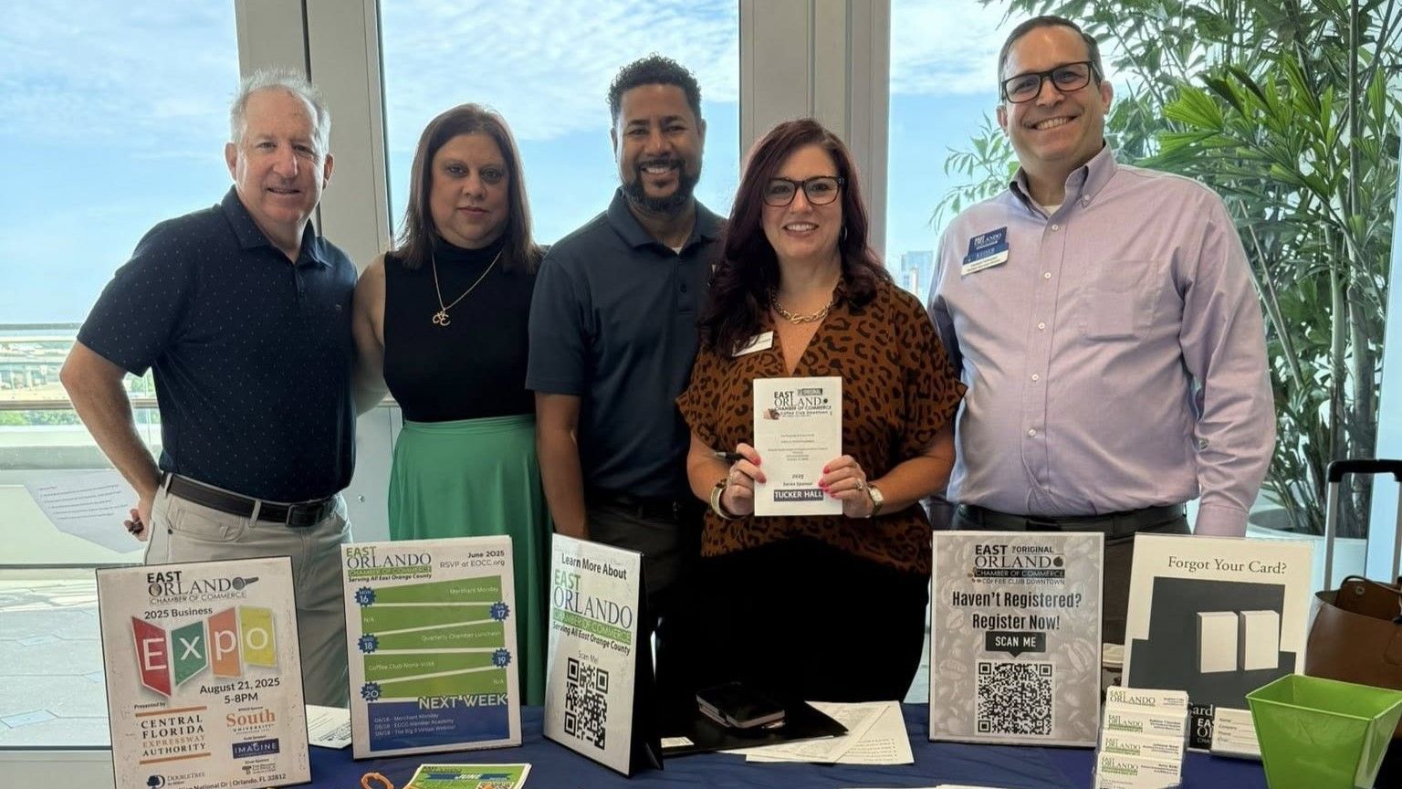 Five people stand at a table with brochures; a window and greenery are behind them.