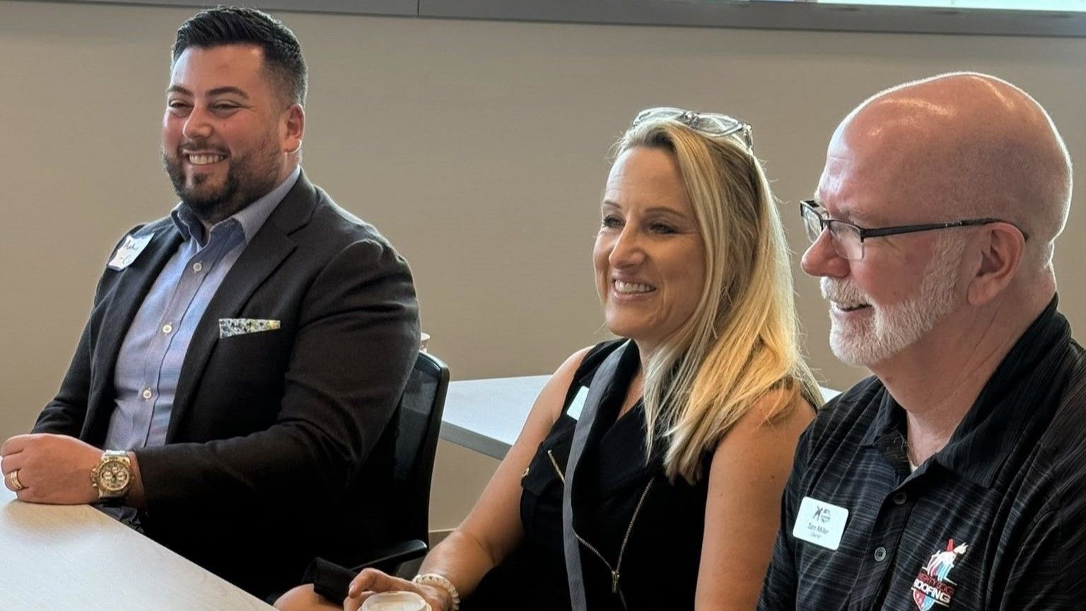 Three people at a table, smiling. Man in suit, woman in black, man with glasses. Neutral setting.