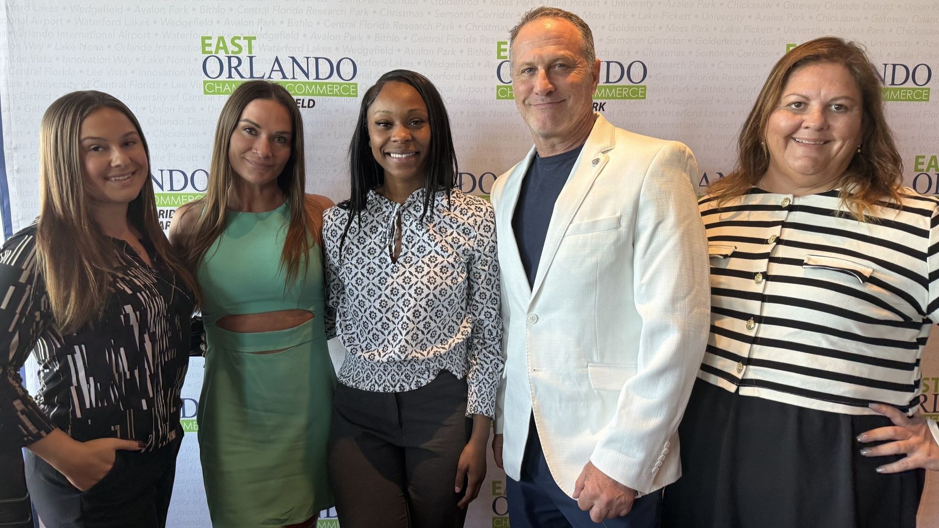 Five people pose for a photo in front of a backdrop with logos. They are smiling.