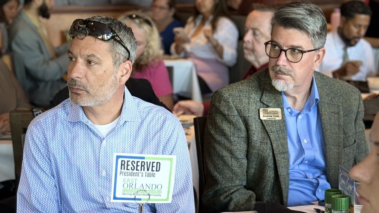 Two men seated at a table, one with sunglasses atop his head. 