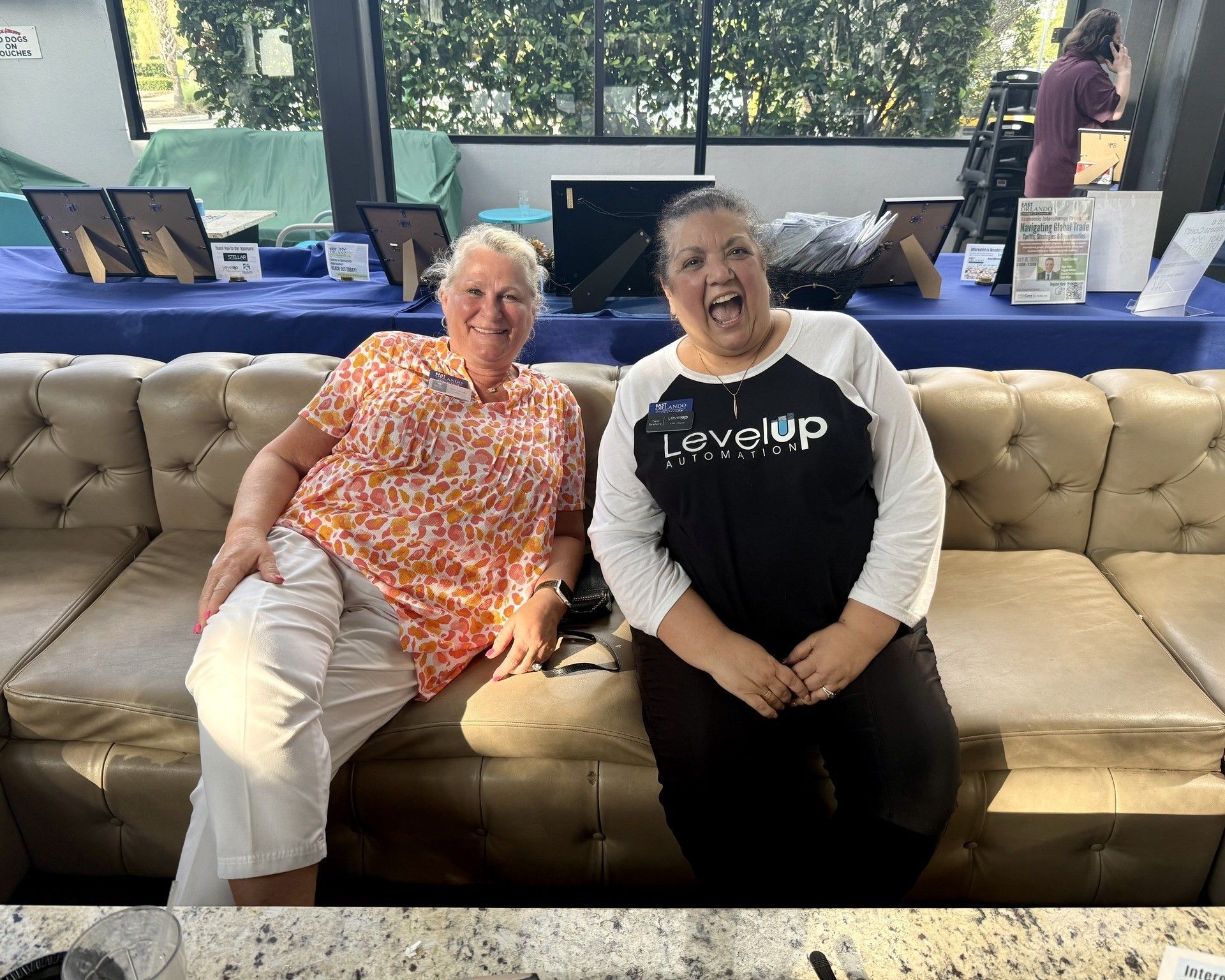 Two women smiling on a brown couch; one wears an orange shirt, the other a Level Up shirt.