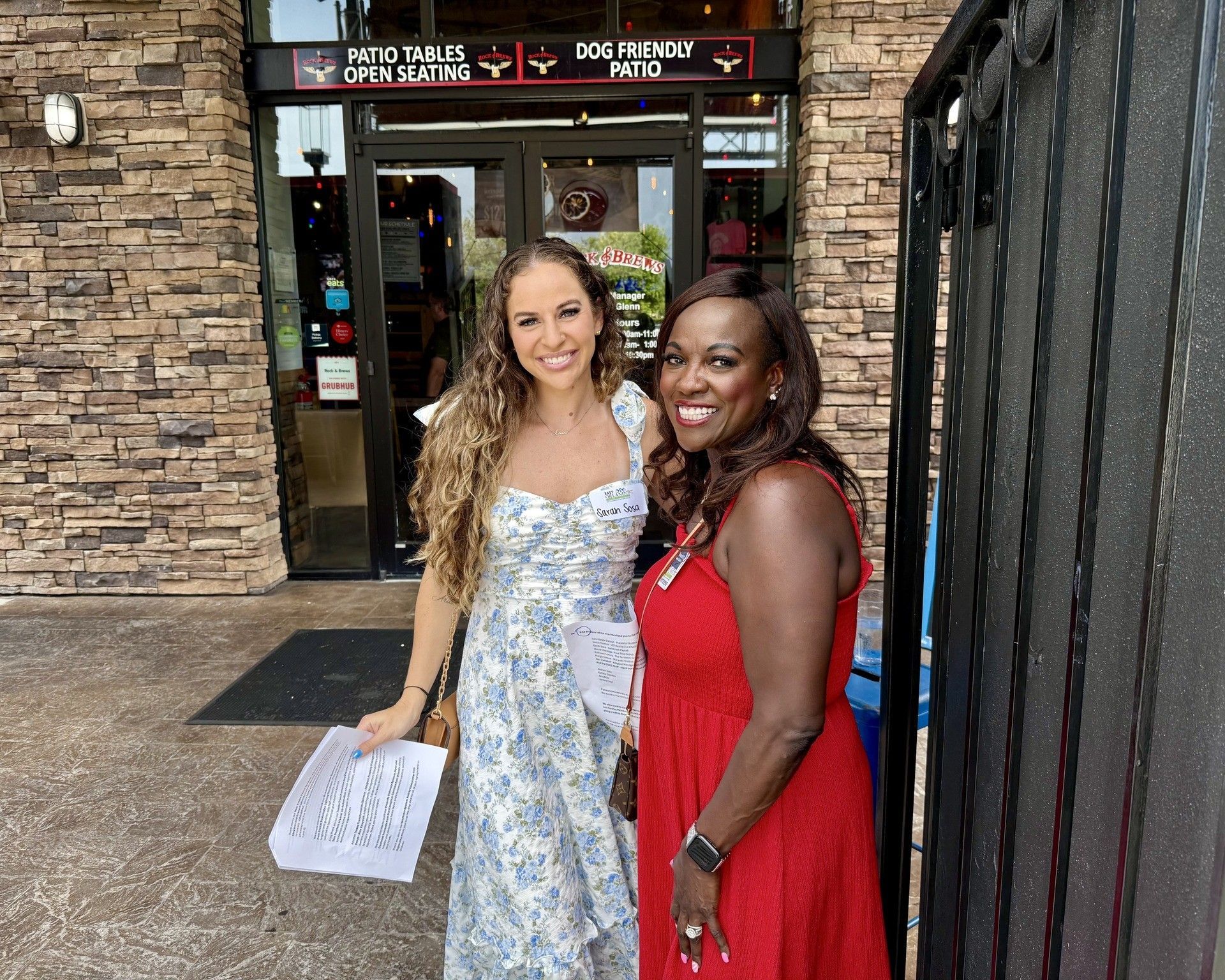 Two women stand outside a restaurant. One wears a floral dress and the other a red dress, smiling.