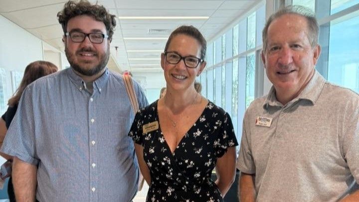 Three people smiling in a bright hallway; a man with glasses and a button-down shirt, a woman with glasses and a floral dress, and a man in a polo shirt.