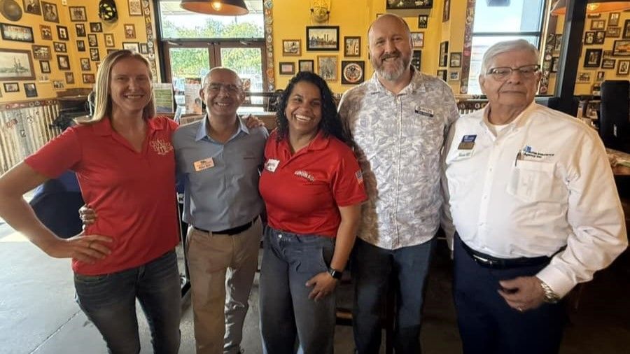 Five people standing closely together inside a restaurant, smiling. Two in red shirts, others in business casual. Yellow walls, framed art.