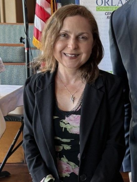 Woman in black blazer, floral top, smiling, standing near an American flag, Orlando Chamber sign.