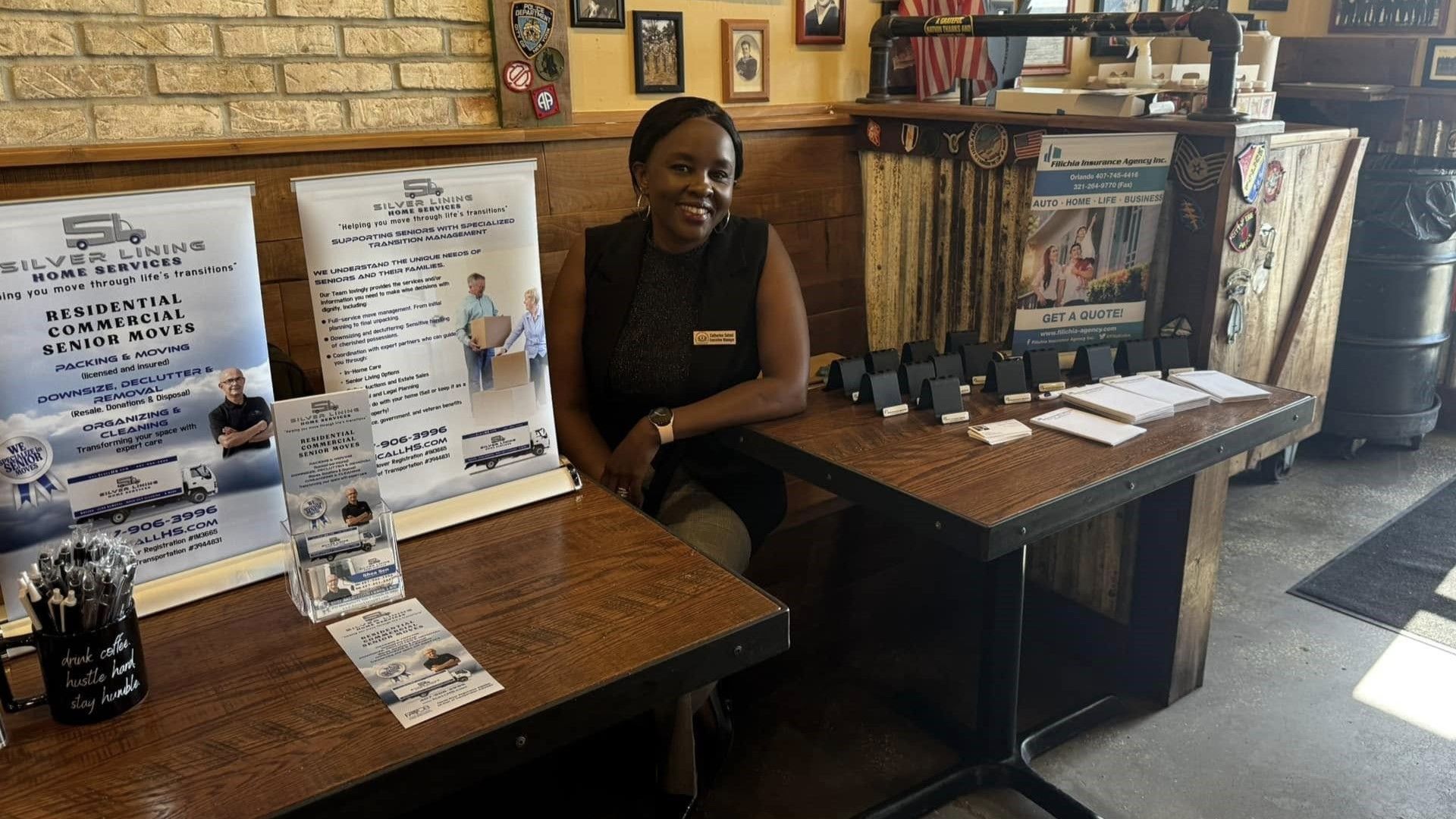 Woman smiling at table with brochures and black boxes in a room with brick wall and decorations.