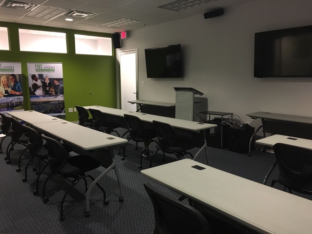 Classroom with tables, chairs, screens, and podium. White tables, black chairs, green wall accent, and blue carpet.