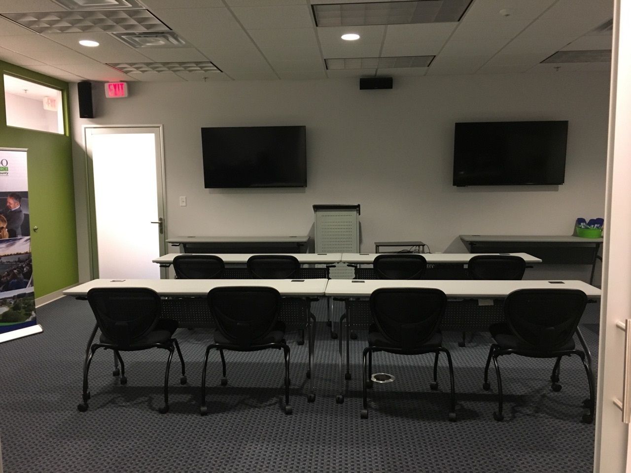 Training room with tables, chairs, two TVs, and a door.