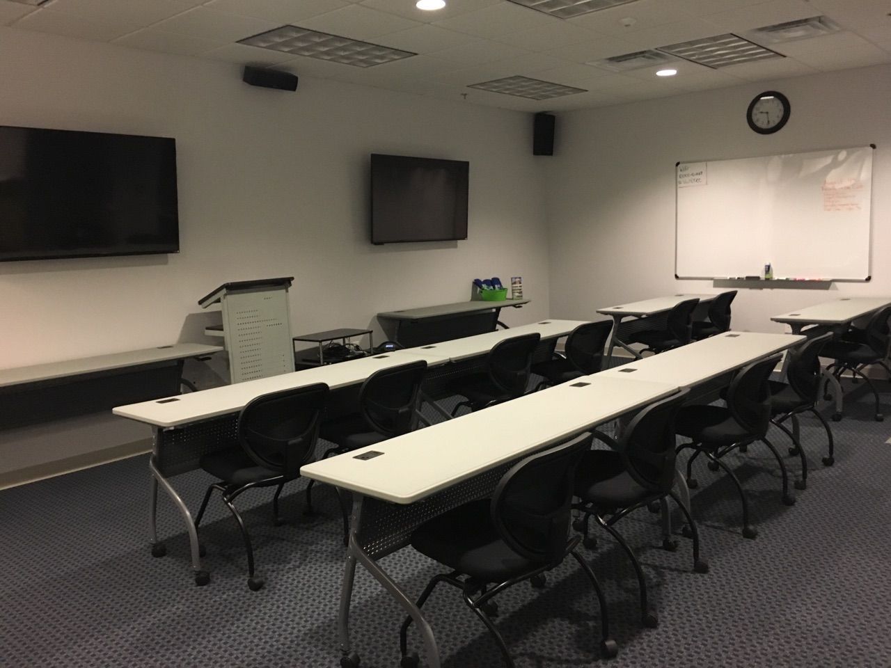 Empty classroom with tables, chairs, two TVs, whiteboard, clock, and sound equipment.