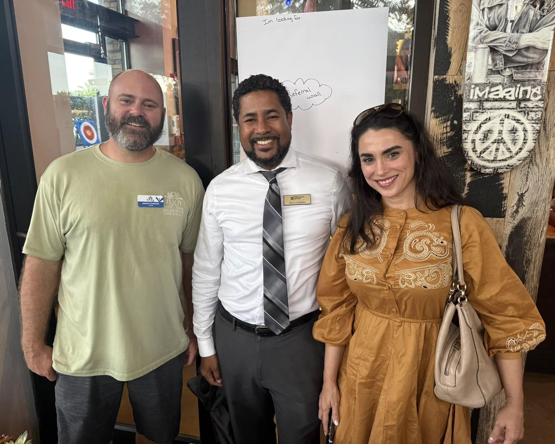 Three people stand indoors: a man in a green shirt, a man in a tie, and a woman in a dress. They are smiling.