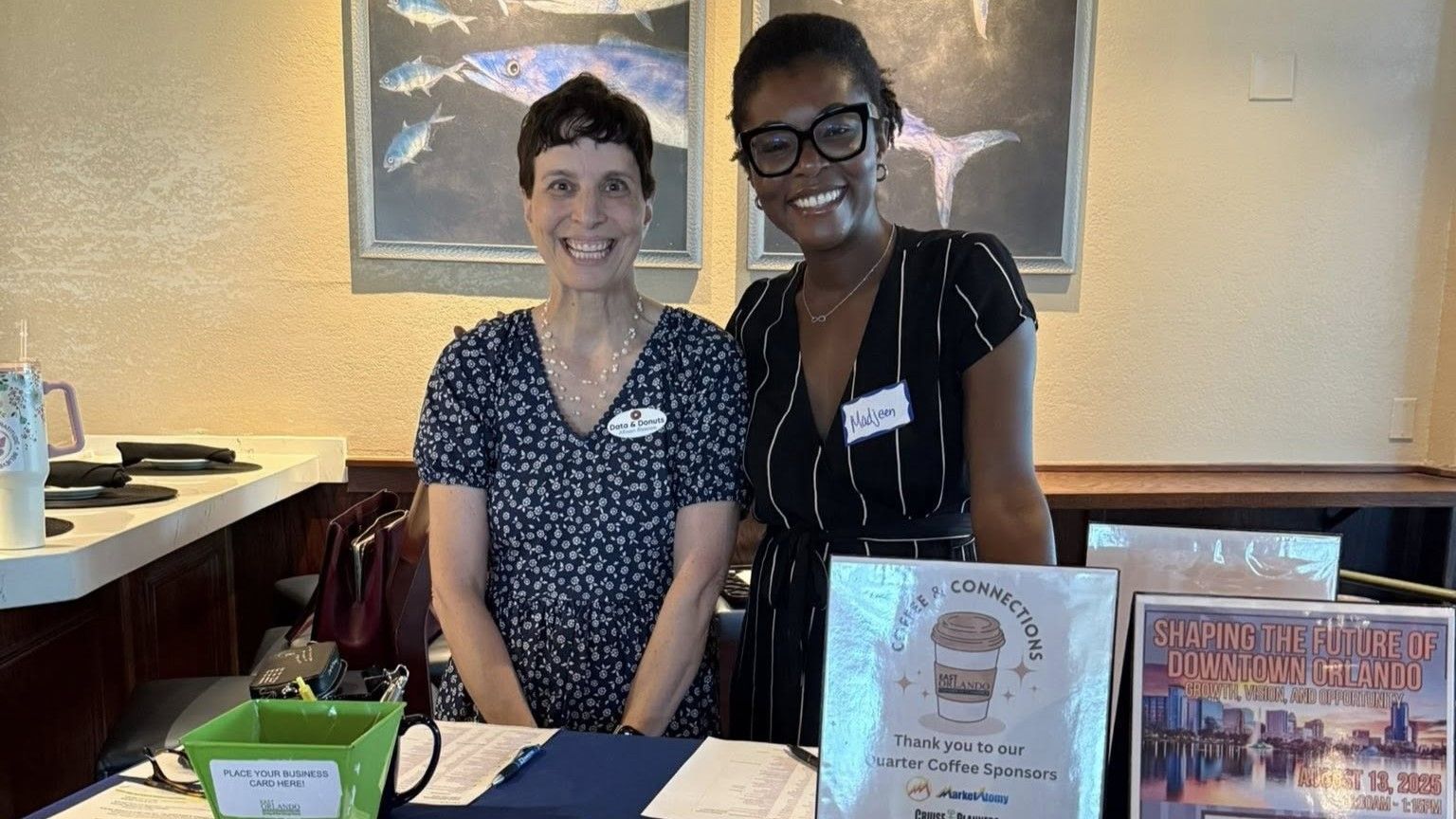 Two women smiling behind a table with promotional materials. Artwork hangs on the wall behind them.