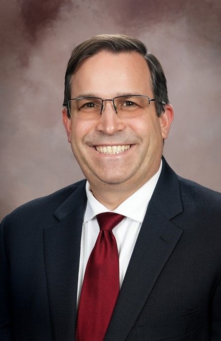 Man in a suit smiles, wearing glasses and a red tie against a blurred background.