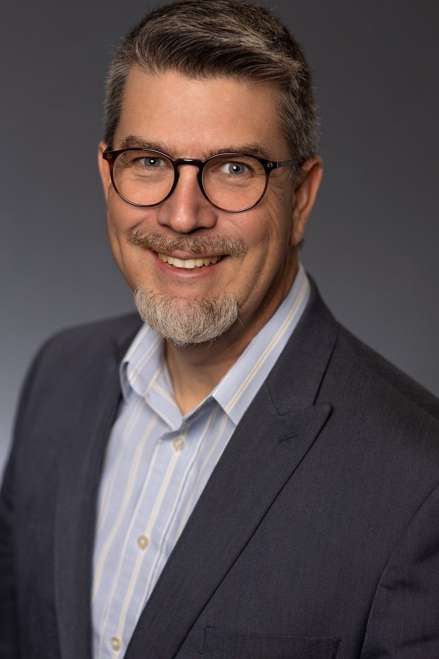 Man with glasses smiling, wearing a blue blazer over a light blue pinstriped shirt; a neutral background.