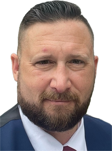 Man with a beard, in a suit, looking at the camera. He has dark hair and a small blemish on his forehead.