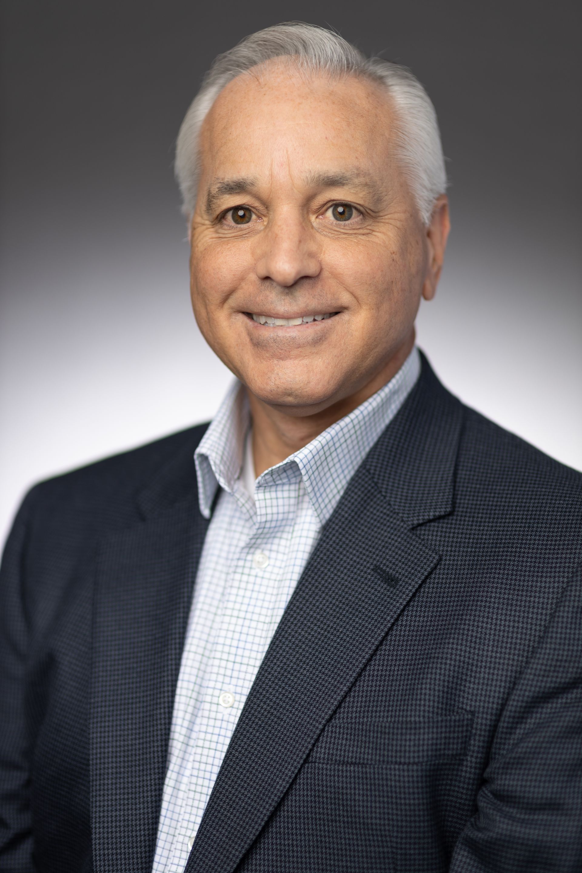 Man in a blue blazer and button-down shirt smiles at the camera. Neutral gray background.