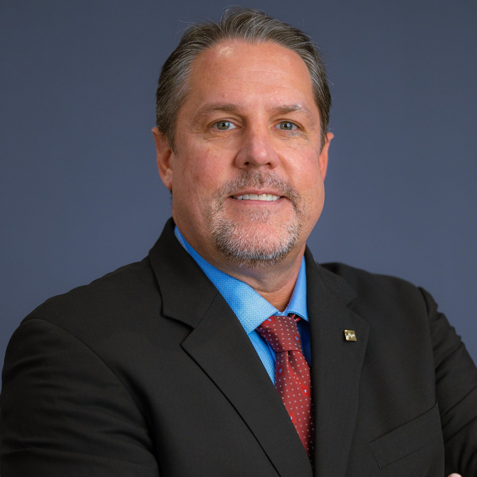 Man with dark hair smiles, wearing a blue dress shirt  red tie and dark blazer; studio headshot.
